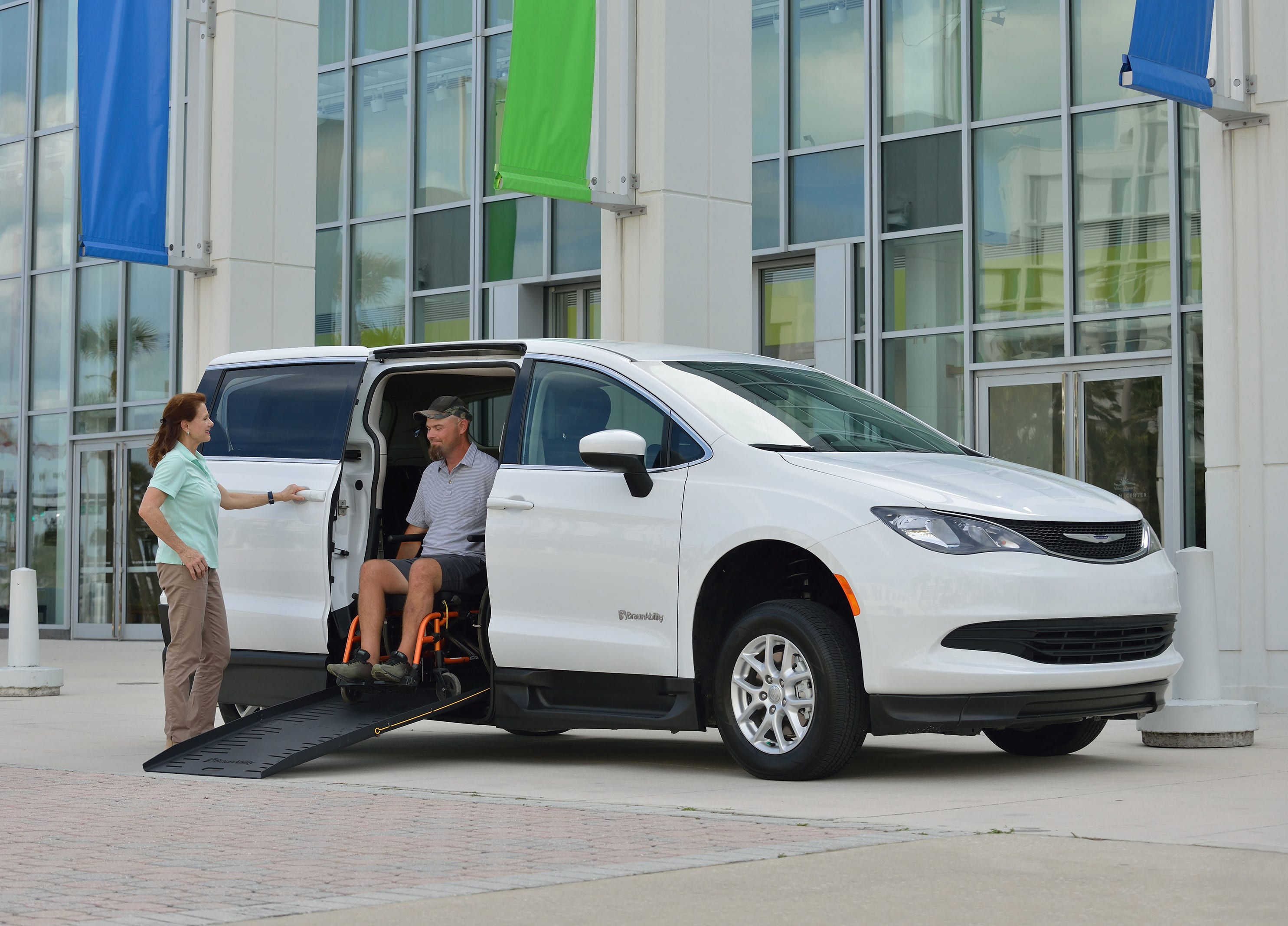 A white Chysler Voyager commercial side entry wheelchair van with a lady operating the vehicle and a man getting ready to go down the ramp